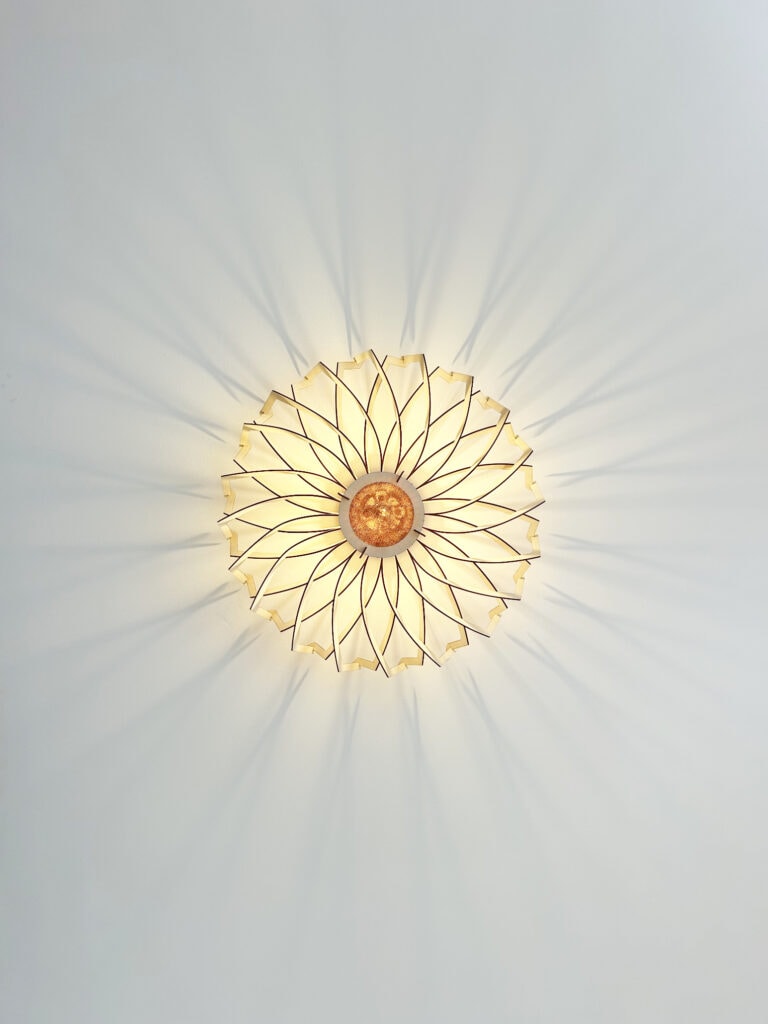 Plafonnier rond en bois découpé modèle Soleil Atelier Sapi fixé au plafond, photographié du dessous, projetant des ombres rayonnantes sur un mur blanc avec corniche moulurée