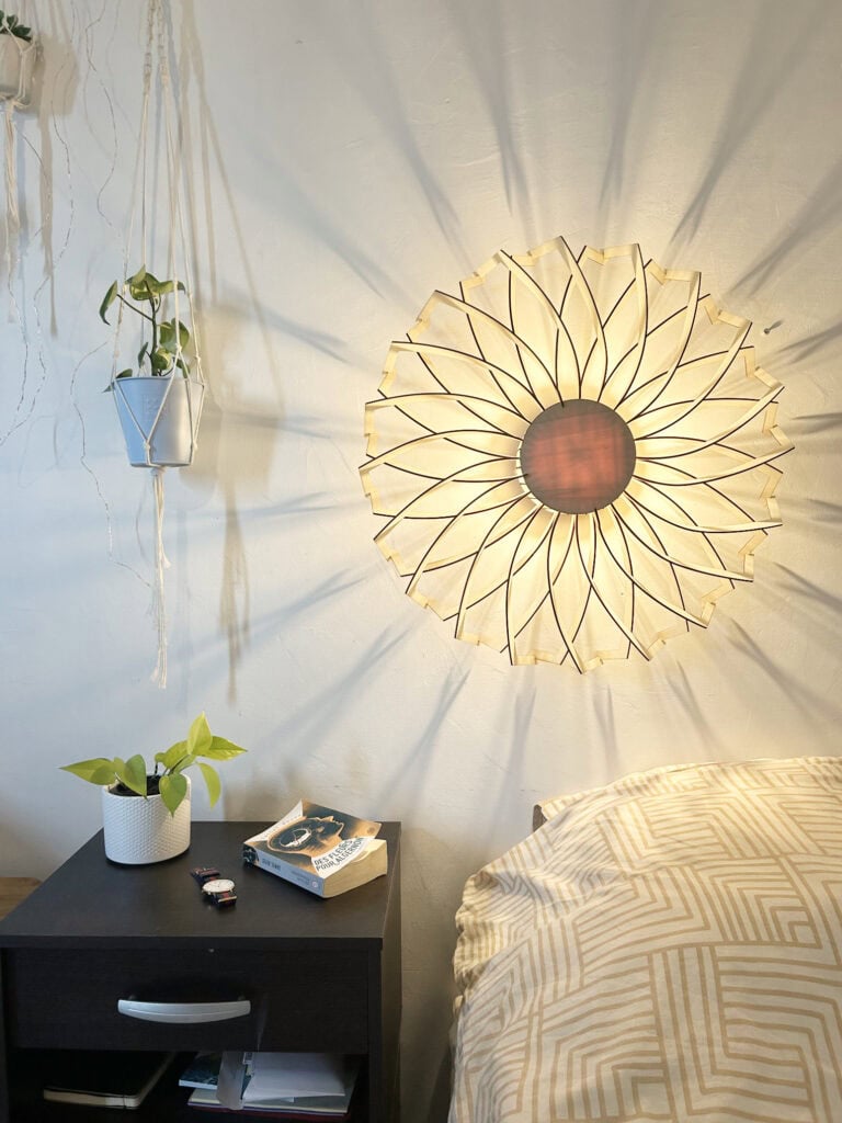 Applique murale modèle Marguerite en bois découpé Atelier Sapi, double rangée de pétales droits formant un mandala fleuri, fixée au-dessus du lit, ombres rayonnantes sur mur blanc, plante suspendue et livre Des Fleurs pour Algernon sur la table de nuit