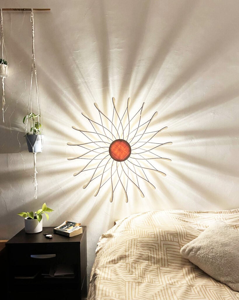 Applique murale modèle Soleil en bois découpé Atelier Sapi fixée au mur au-dessus d'un lit, lumière chaude dorée, ombres rayonnantes sur mur blanc, plantes suspendues et table de nuit noire avec livre