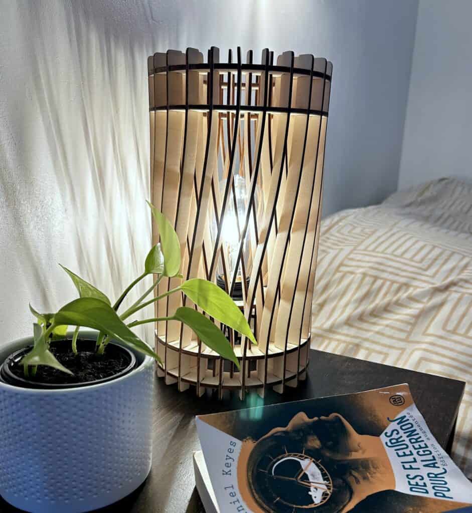 Lampe de chevet en bois découpé modèle torsadé Atelier Sapi posée sur une table de nuit, entourée d'une plante et d'un livre, en lumière naturelle