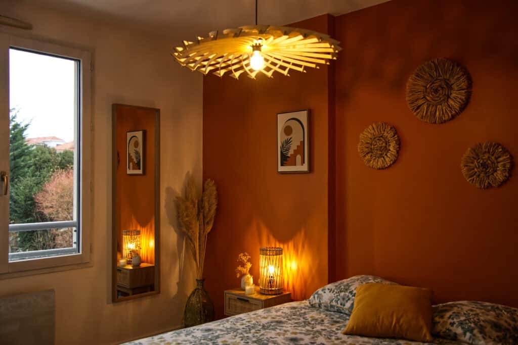 Chambre décorée avec une suspension soleil en bois Atelier Sapi au plafond et une lampe cylindrique sur la table de chevet, mur terracotta, lit avec coussin moutarde