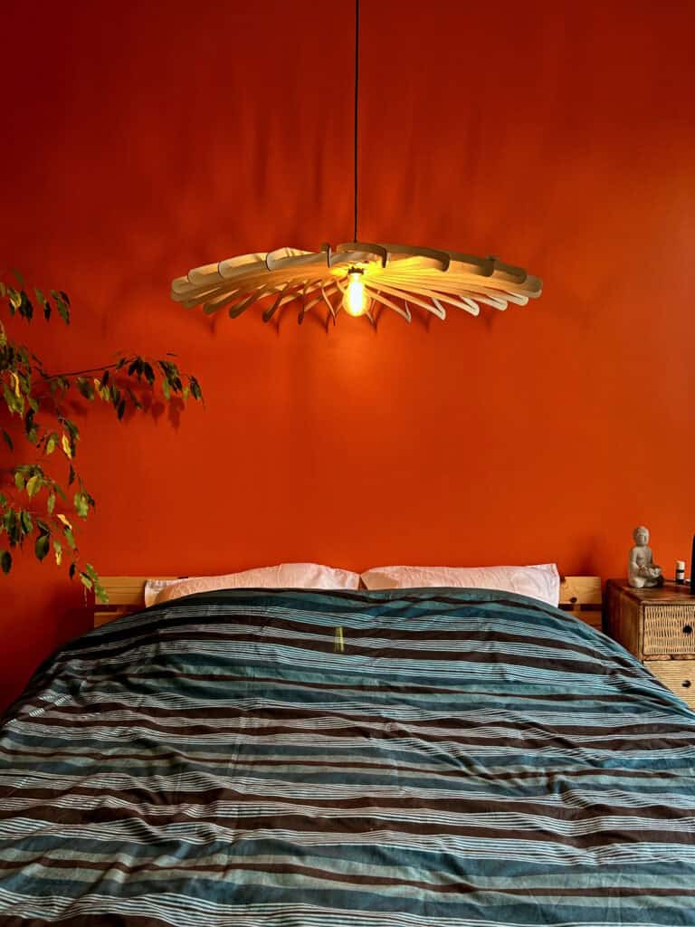 Suspension en bois découpé Atelier Sapi modèle Chapeau Chinois au-dessus d'un lit double, mur rouge vif, plante verte et statuette Bouddha