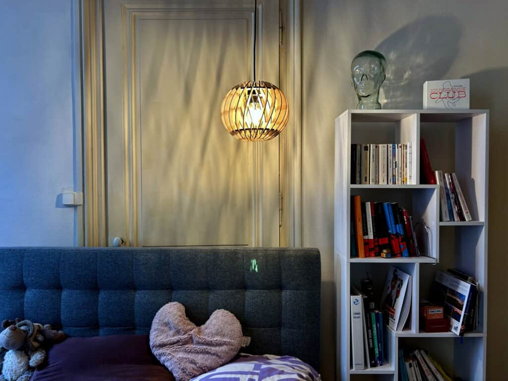 Suspension sphérique en bois découpé Atelier Sapi accrochée dans une chambre avec porte ancienne en bois, bibliothèque blanche et tête de lit grise