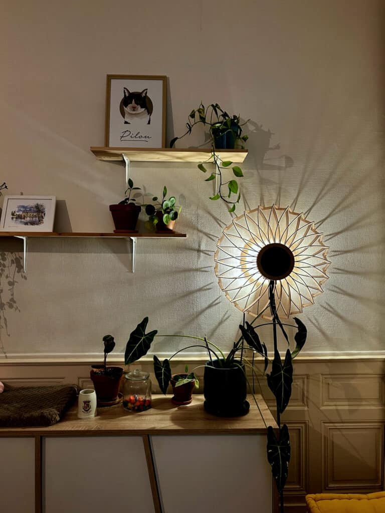 Lampadaire sur pied modèle Soleil en bois découpé Atelier Sapi allumé le soir dans un salon, projetant des ombres en rayons de soleil sur un mur blanc avec moulures