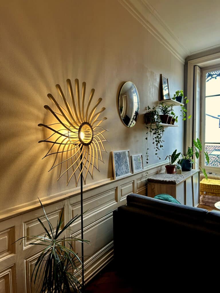 Lampadaire sur pied modèle Soleil Atelier Sapi allumé dans un salon haussmannien avec moulures blanches, miroir ovale, plantes vertes et fenêtre ouverte