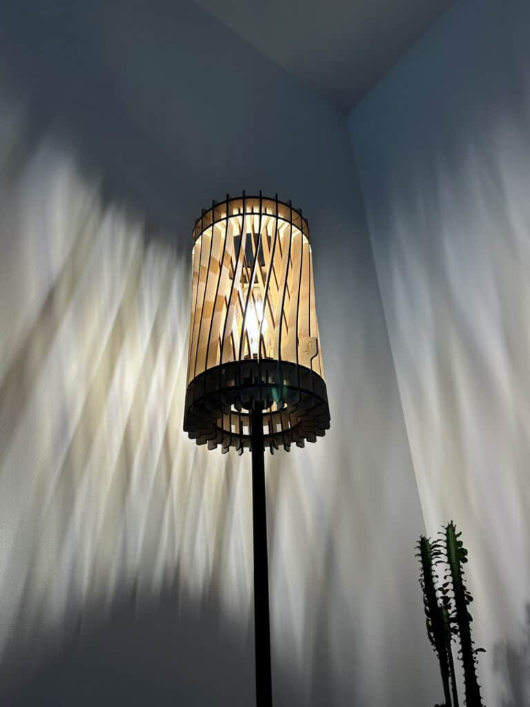 Lampadaire sur pied modèle Torsadé cylindrique en bois découpé Atelier Sapi, photographié en contre-plongée, projetant des ombres verticales ondulantes sur un mur blanc, cactus au premier plan