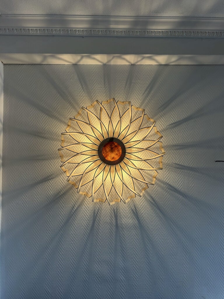 Plafonnier rond en bois découpé modèle Soleil Atelier Sapi fixé au plafond, photographié du dessous, projetant des ombres rayonnantes sur un mur blanc avec corniche moulurée