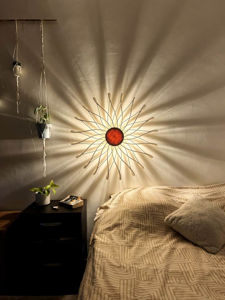 Applique murale modèle Soleil en bois découpé Atelier Sapi fixée au mur au-dessus d'un lit, lumière chaude dorée, ombres rayonnantes sur mur blanc, plantes suspendues et table de nuit noire avec livre