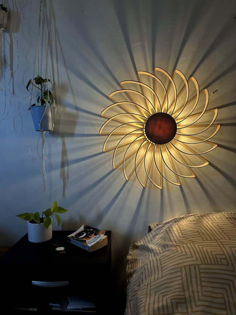 Applique murale modèle Hélios en bois découpé Atelier Sapi, pétales en spirale, fixée au mur au-dessus d'une tête de lit, lumière chaude dorée, ombres spiralées rayonnantes sur mur blanc, plante suspendue et table de nuit noire avec livre Des Fleurs pour Algernon