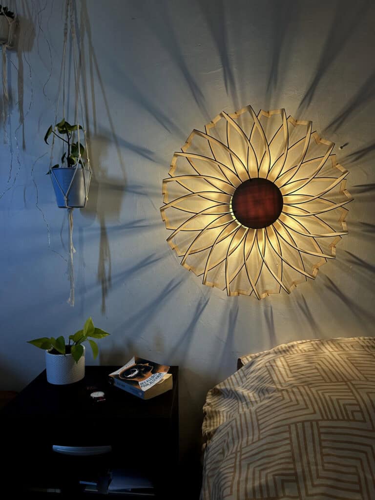 Applique murale modèle Marguerite en bois découpé Atelier Sapi, double rangée de pétales droits formant un mandala fleuri, fixée au-dessus du lit, ombres rayonnantes sur mur blanc, plante suspendue et livre Des Fleurs pour Algernon sur la table de nuit