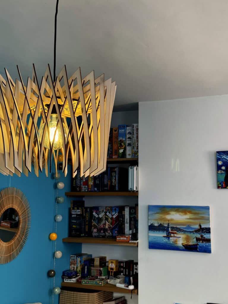 Suspension modèle Couronne en bois découpé Atelier Sapi, lamelles verticales allongées formant un cylindre ajouré, ampoule Edison visible, accrochée dans un salon au mur bleu canard, étagères de jeux de société et tableau Nuit Étoilée en fond