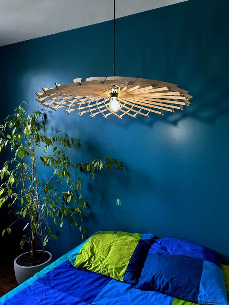 Suspension grand format modèle Galaxie Atelier Sapi, lamelles en spirale formant un disque plat de grand diamètre, ampoule centrale allumée, accrochée au-dessus d'un lit devant un mur bleu pétrole, linge de lit bleu et vert, ficus