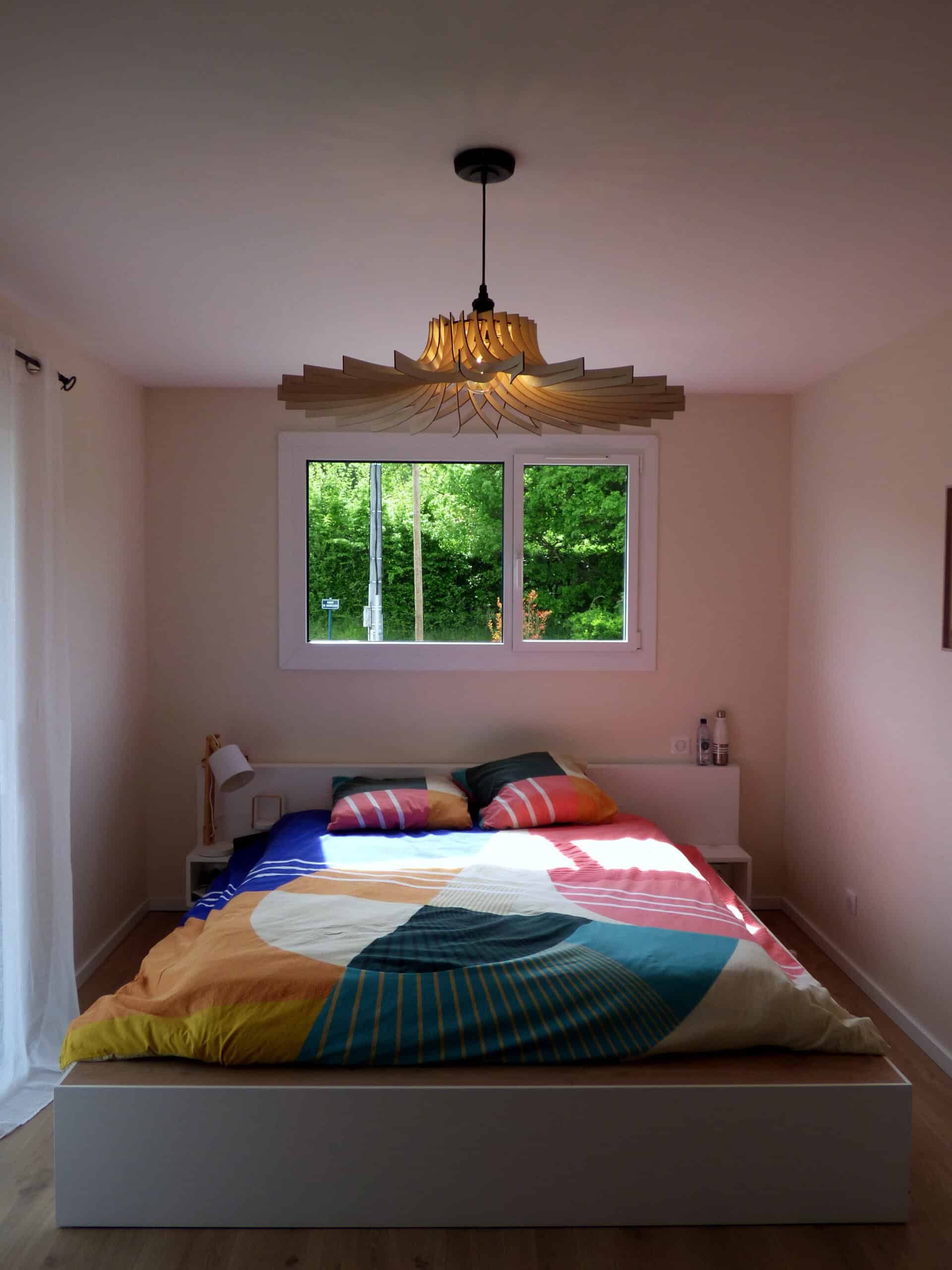 Suspension modèle Éclat Atelier Sapi en bois découpé, grandes lamelles plates rayonnantes formant un disque ouvert, accrochée au-dessus d'un lit blanc avec housse colorée, chambre claire ouverte sur la nature par une grande fenêtre