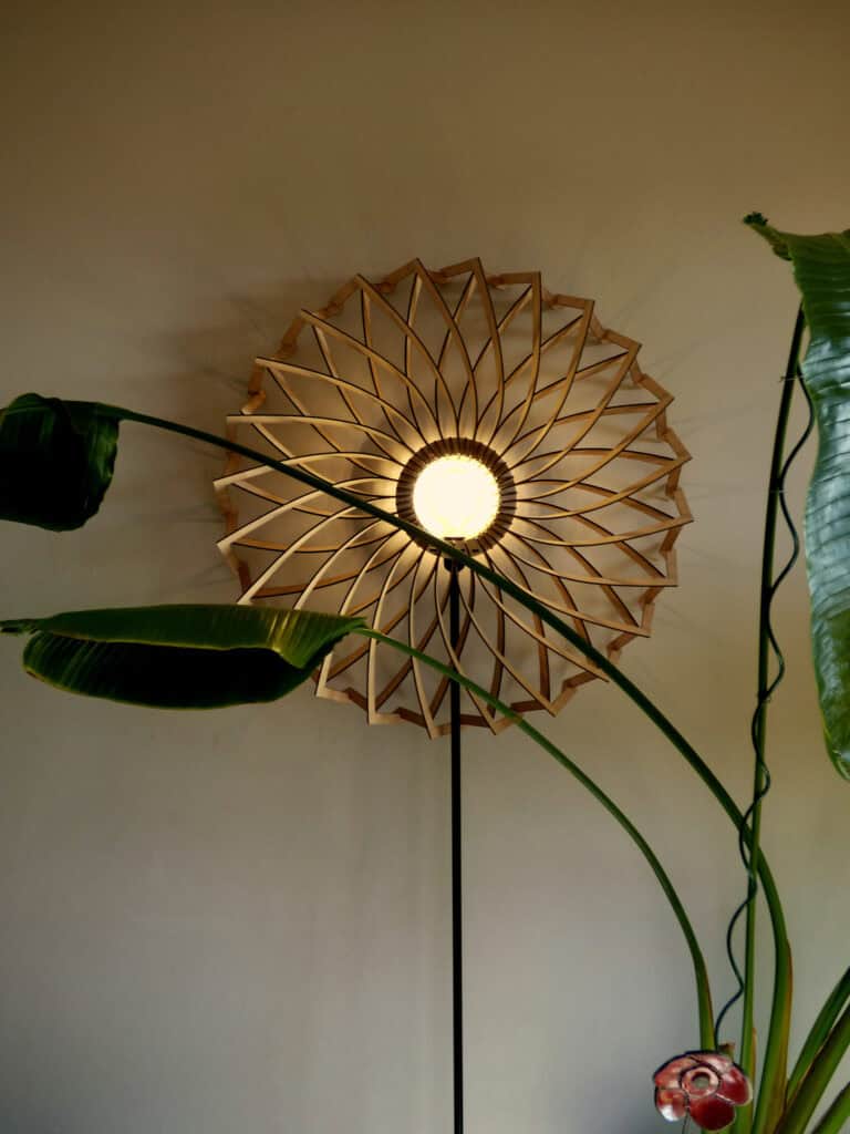 Lampadaire modèle Marguerite Atelier Sapi, grand abat-jour plat en bois découpé motif mandala à double rangée de pétales, pied noir fin, entouré de feuilles de bananier et strelitzia