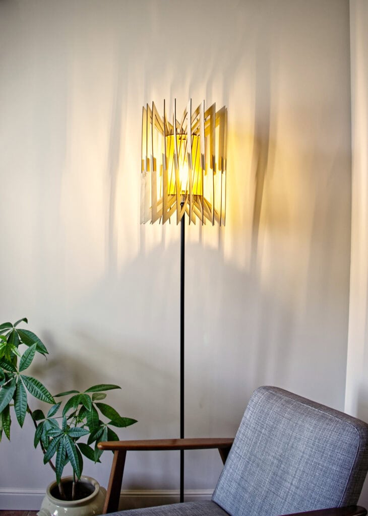 Lampadaire Léon allumé à côté d'un fauteuil gris moderne, projetant des ombres graphiques sur le mur.