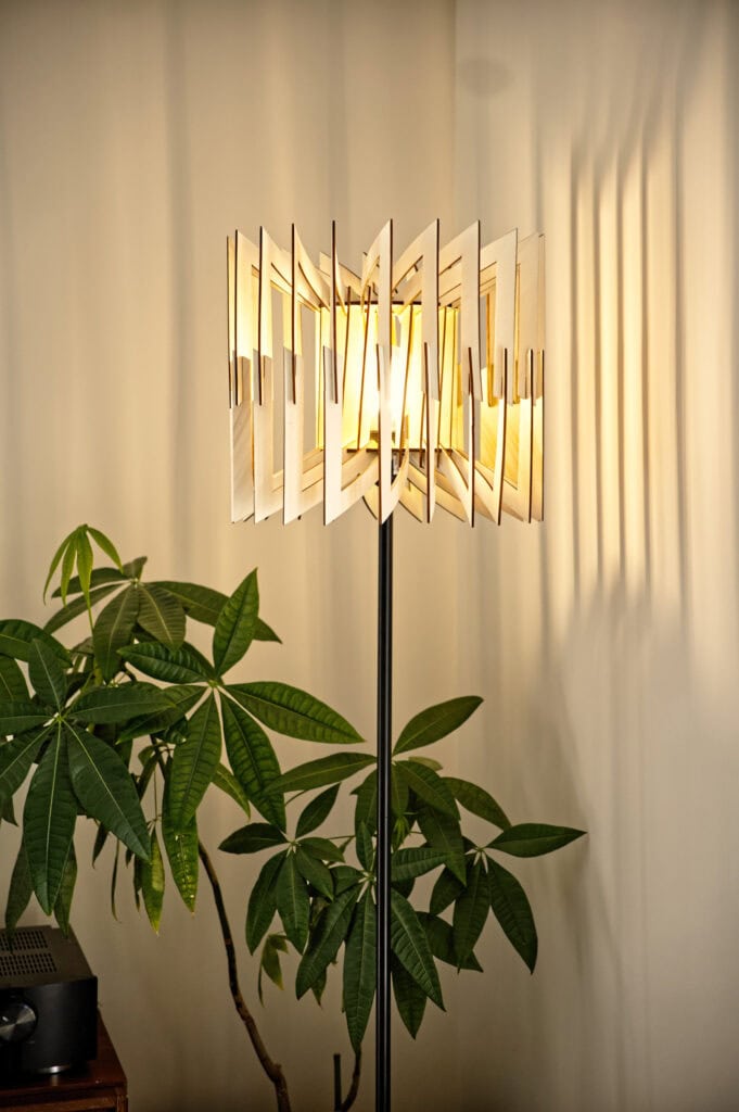 Le lampadaire géométrique Léon allumé contrastant avec les grandes feuilles d'une plante verte d'intérieur.