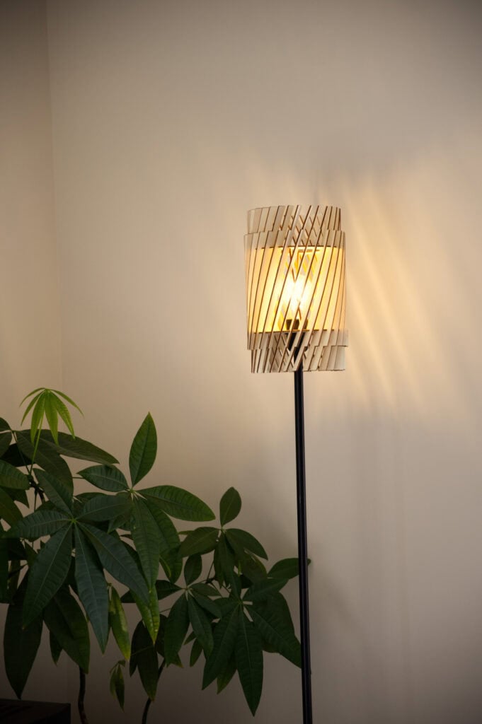 Lampadaire en bois Alice allumé, feuillage d'une plante au premier plan